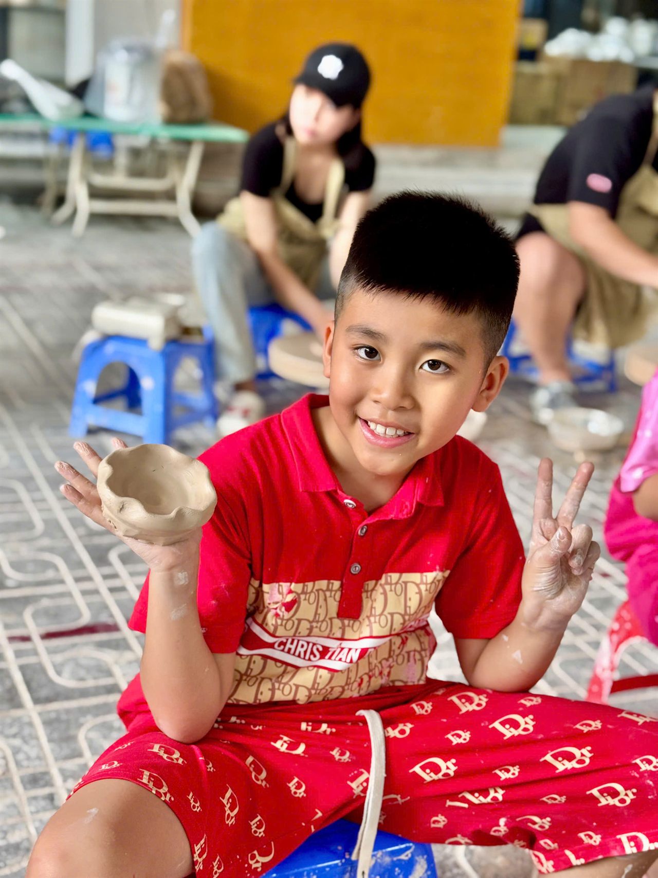 Bé trai cười, giơ chiếc bát gốm tự nặn tại xưởng gốm
