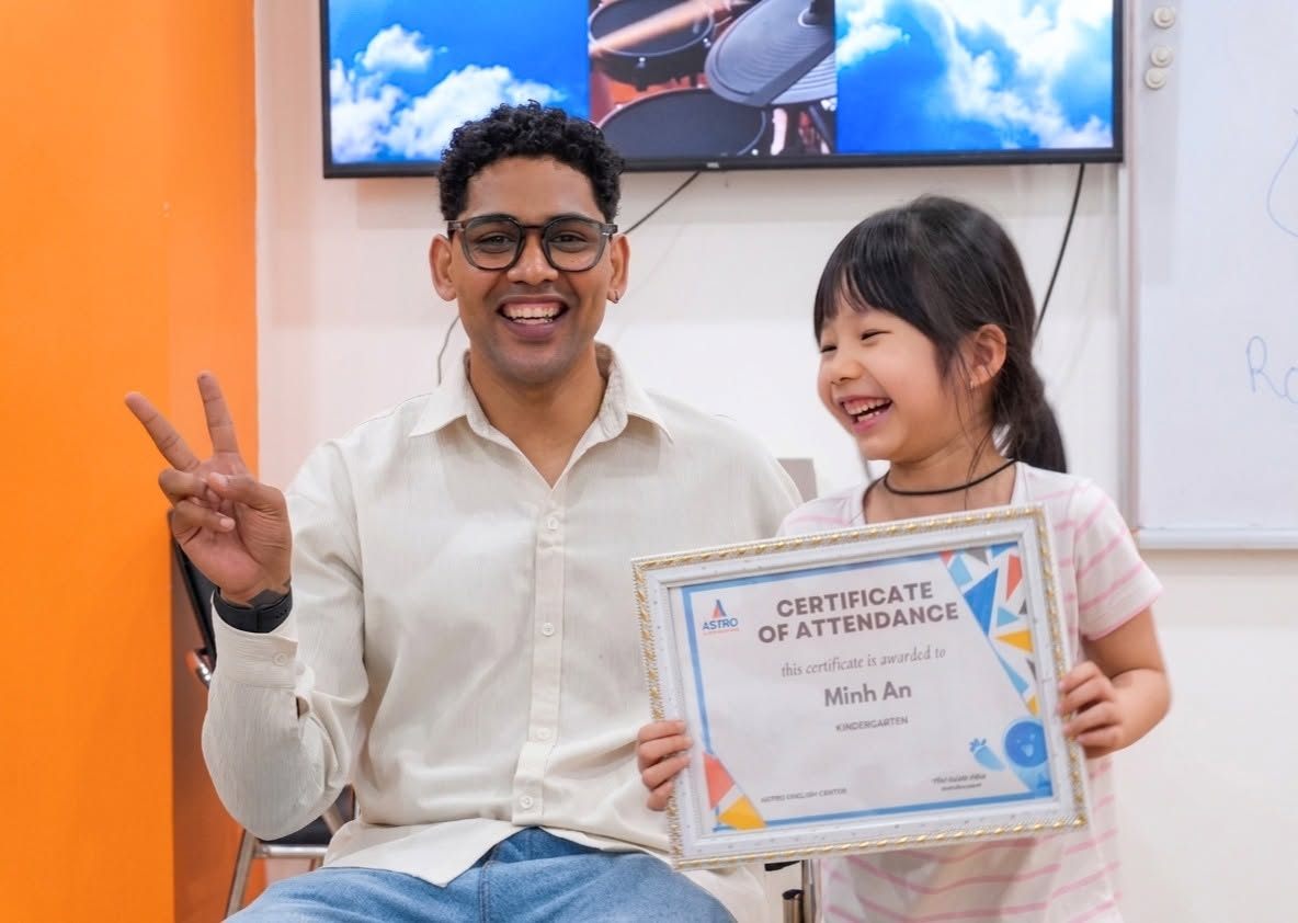 Bé Minh An cầm chứng nhận của Astro English Center, đứng cạnh giáo viên