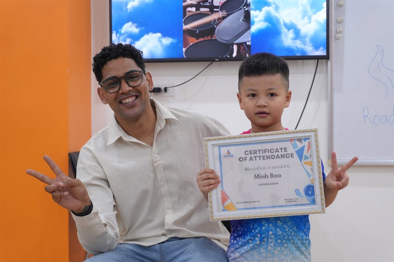 Bé Minh Bao cầm chứng nhận của Astro English Center, ngồi cạnh giáo viên trong lớp