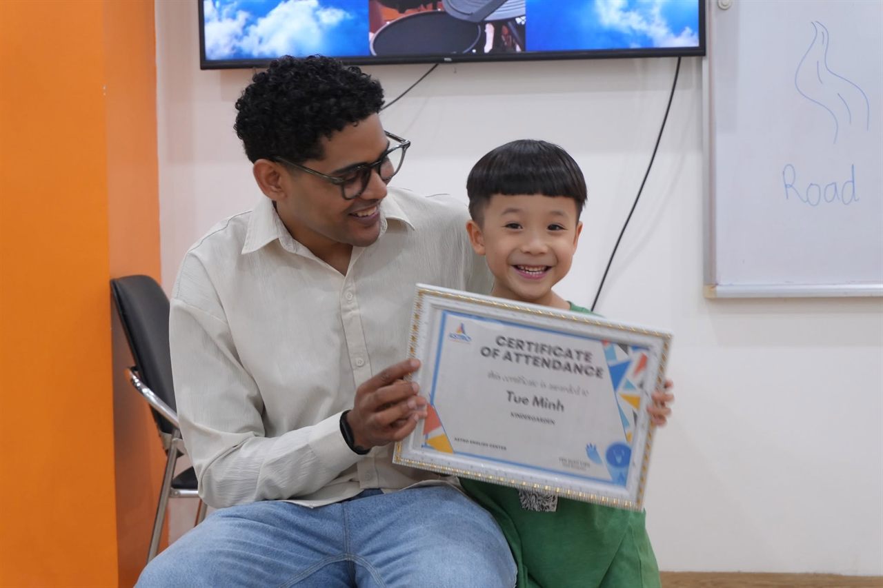 Bé Tue Minh cười tươi, cùng giáo viên cầm chứng nhận của Astro English Center